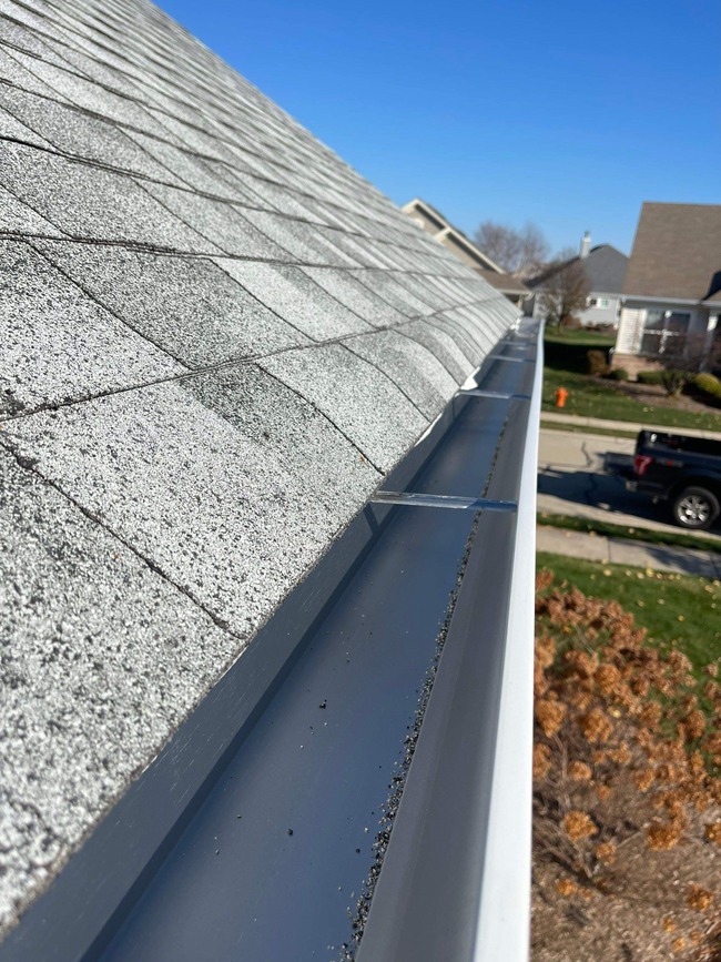 Close-up, angled view of a newly installed, large-capacity seamless gutter system on a residential roof with light gray asphalt shingles. The professional installation highlights the clean, wide gutter securely fastened below the roofline.